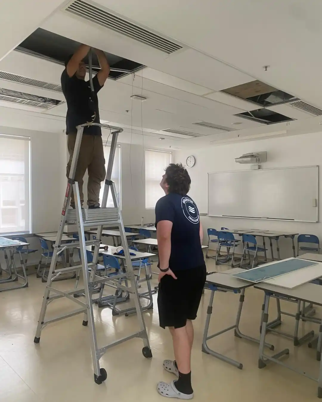 An AirconPro technician on a ladder servicing a ceiling unit while an apprentice observes from below